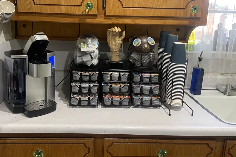 A coffee maker beside rows of coffee and tea pods and stacks of cups.