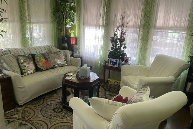 The counseling room: a white sofa and two armchairs around a small table, by tall windows with sheer green curtains and plants.